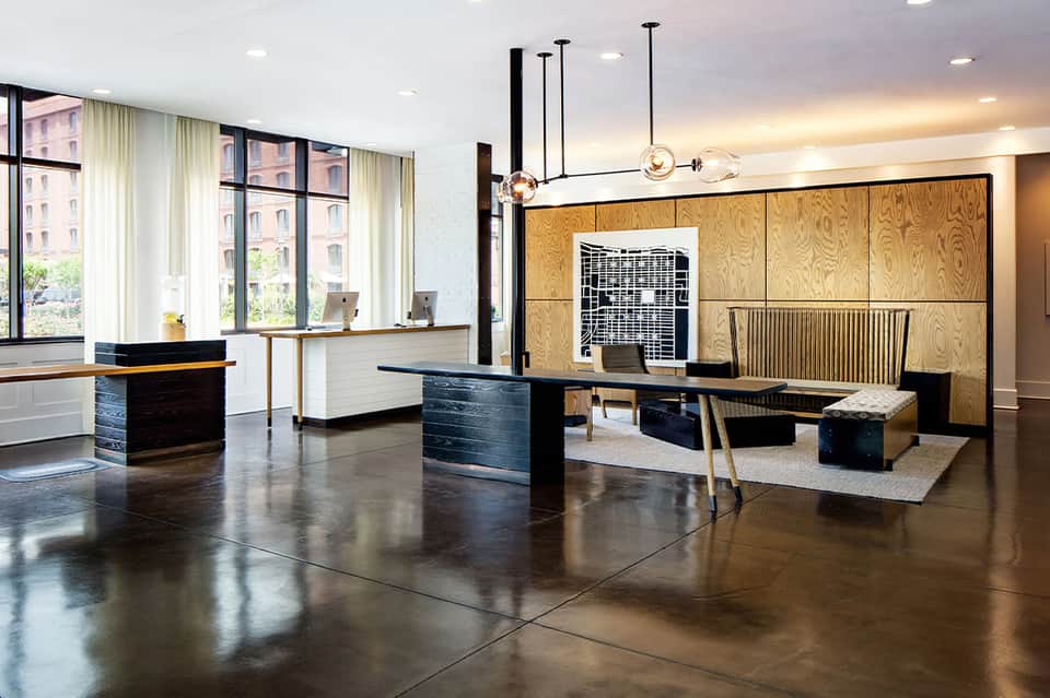 Modern hotel lobby with polished concrete floors, black desks, wood paneling, pendant lighting, and tall windows