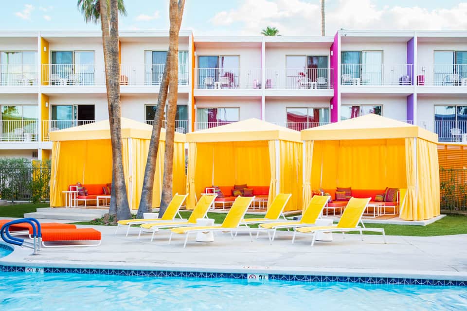 Colorful resort pool area with yellow cabanas, lounge chairs, and palm trees by the water