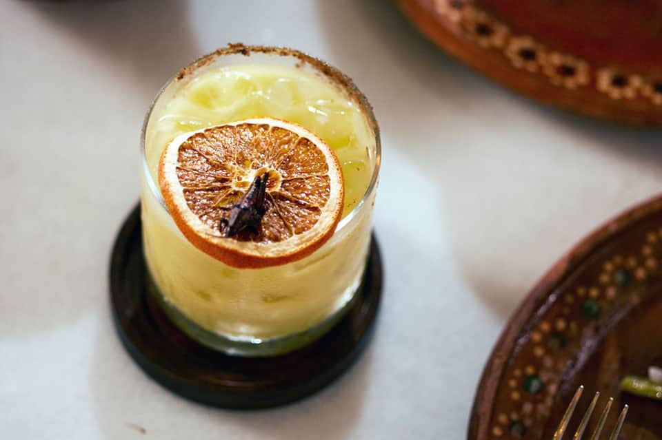 Craft cocktail with orange slice garnish on dark coaster with appetizers