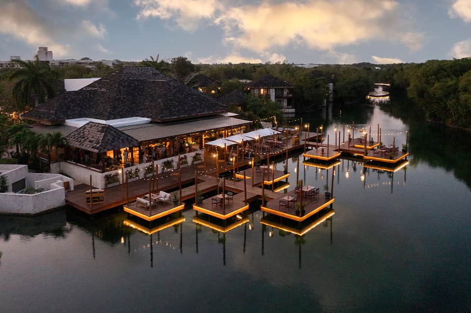 Waterfront dining area at dusk with illuminated floating platforms, dark roof structure, and trees