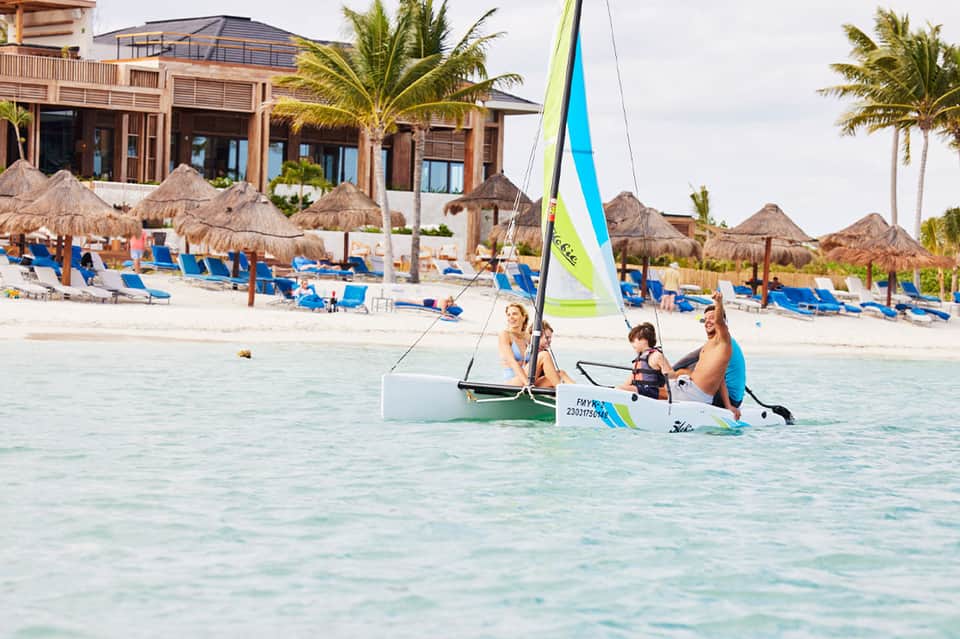Three people sailing in turquoise water with beachfront resort and umbrellas in background