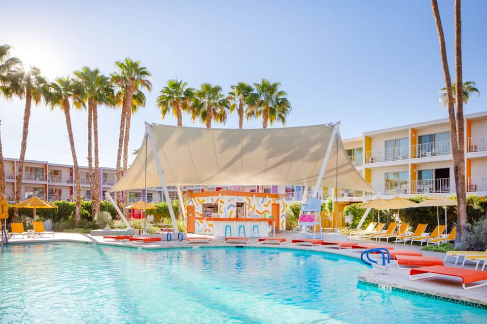 Colorful poolside lounge area with large sail shade, orange bar, yellow loungers, and palm trees