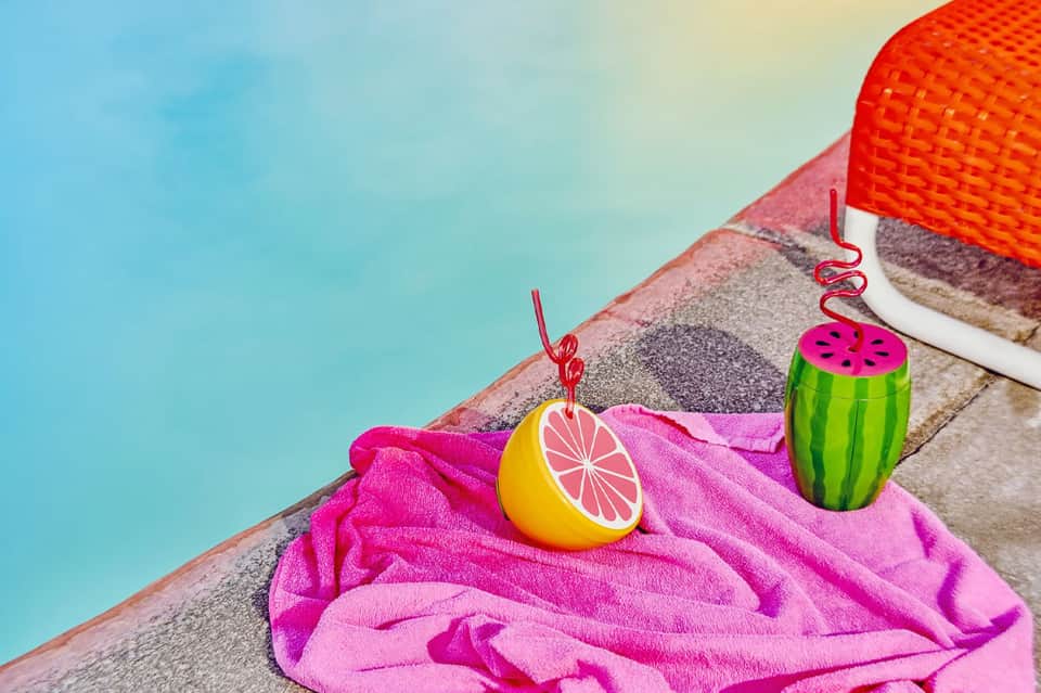 Colorful pool accessories including pink towel, lemon and watermelon drink charms on pool deck