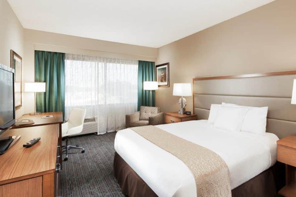 Hotel bedroom with queen bed, desk, green curtains, flat-screen TV, and neutral color palette.