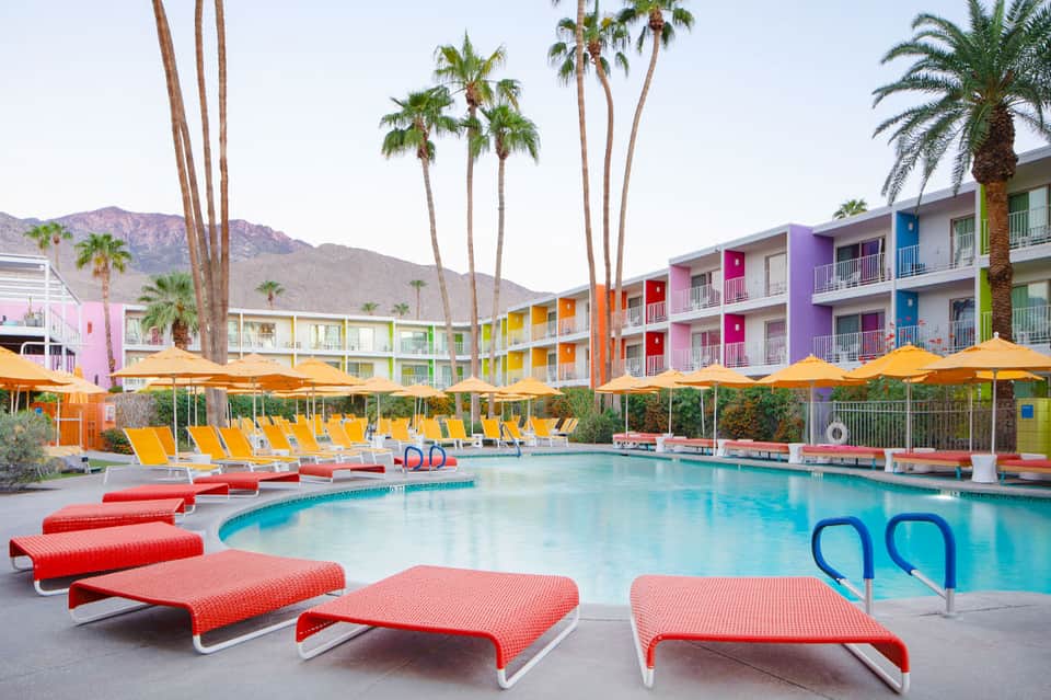 Vibrant resort pool area with colorful mid-century modern buildings, yellow umbrellas, red lounge chairs, and palm trees