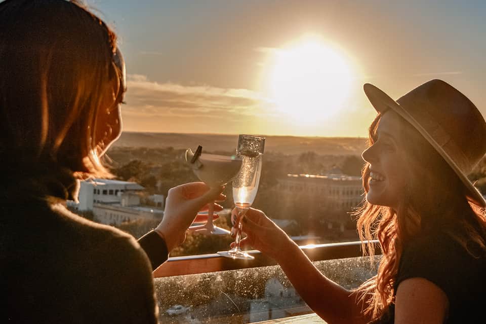 Two women toasting champagne glasses at sunset overlooking coastal city landscape