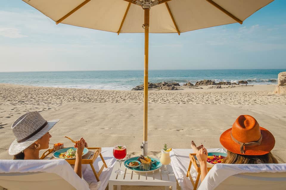 Two women dining under beige umbrella on sandy beach with turquoise ocean and rocky coastline backdrop
