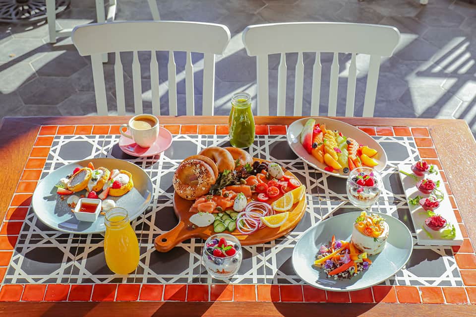 Outdoor dining table with colorful breakfast spread, fresh juices, and pastries on terracotta tile patio