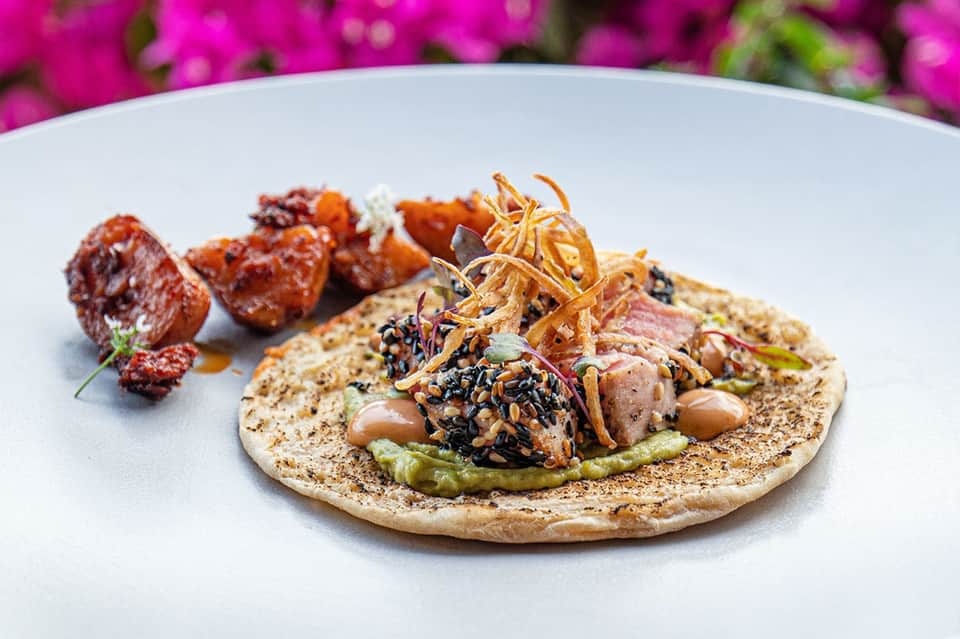 Gourmet plated dish with seared meat, vegetables, black seeds, and magenta bougainvillea flowers backdrop