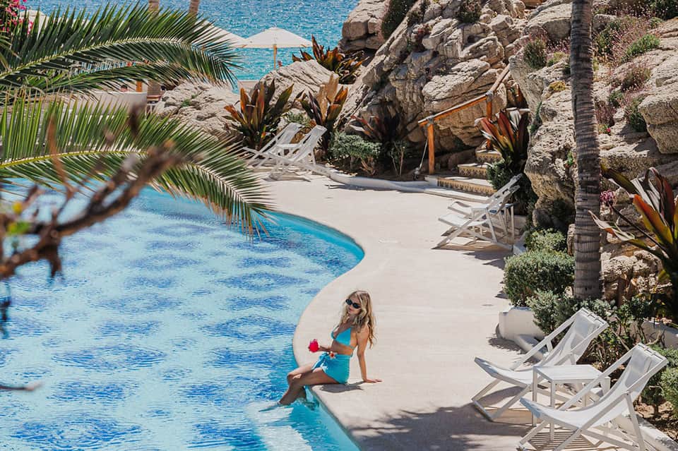Resort guest in turquoise swimsuit sitting at edge of curved pool surrounded by palm trees