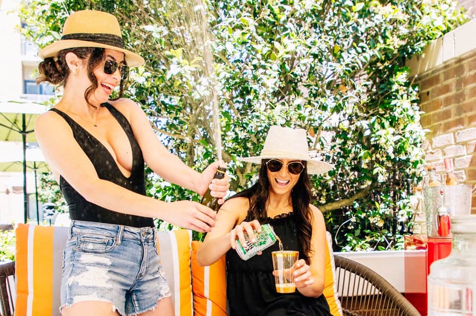 Two women socializing poolside wearing hats and sunglasses with ivy-covered wall backdrop
