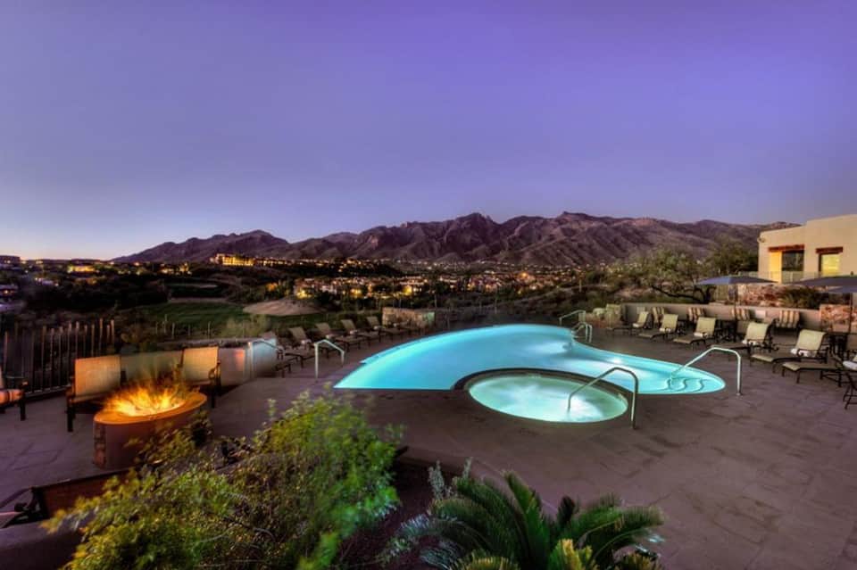 Illuminated pool at dusk with spa tub, firepit, lounge chairs, and mountain views