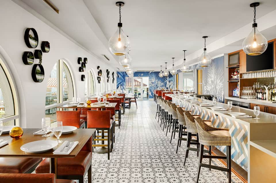 Upscale restaurant with white walls, pendant lights, wooden tables, orange seating, and ocean views through arched windows
