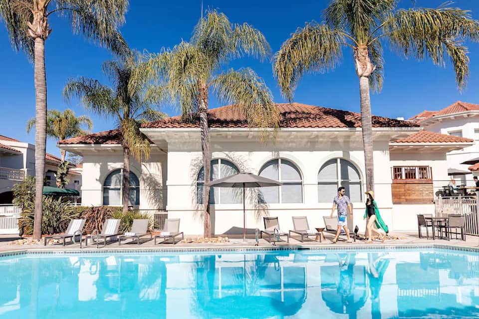 Resort pool with palm trees, white cabana building, and guests walking along pool deck
