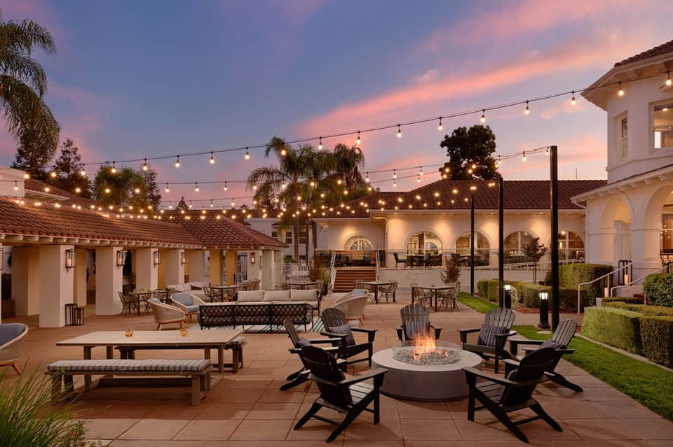Resort patio at twilight with firepit, lounge seating, string lights, and white Spanish architecture
