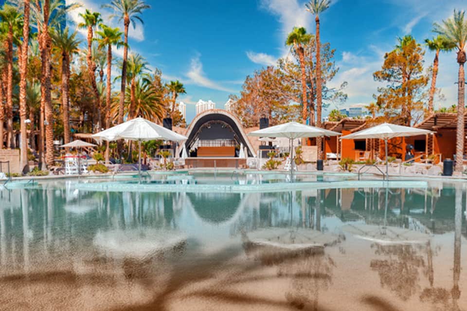 Contemporary resort pool with palm trees, white umbrellas, orange building, and urban skyline