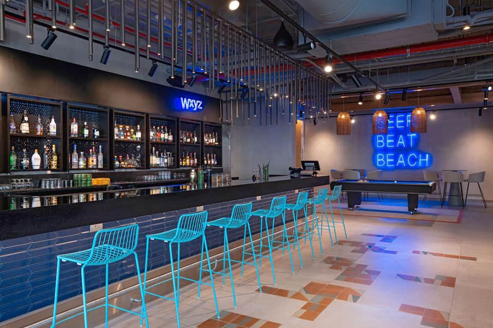 Modern hotel bar interior with blue neon sign reading 'BEAT BEACH' and turquoise bar seating