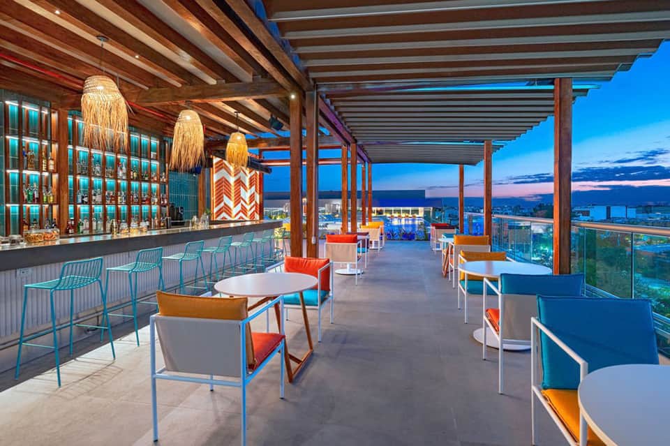 Upscale rooftop bar at dusk with colorful seating, wooden beams, city skyline views at twilight