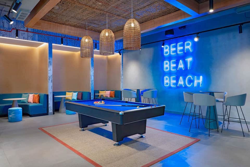 Modern game room with neon 'Beer Beat Beach' sign, blue billiards table, and colorful seating