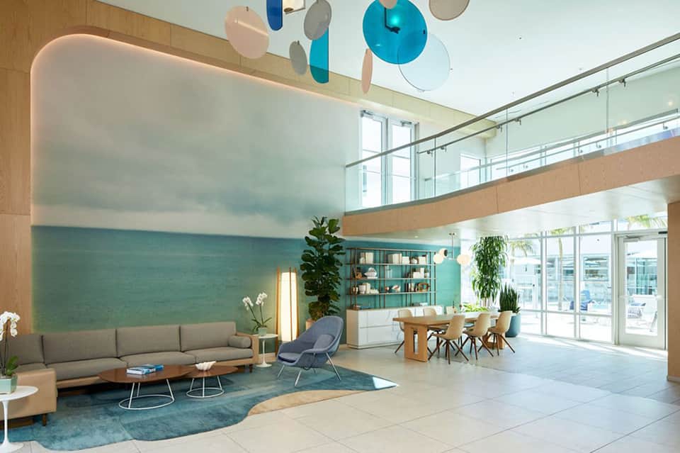 Modern hotel lobby with high ceilings, ocean mural, gray sofa, dining area, and glass railings