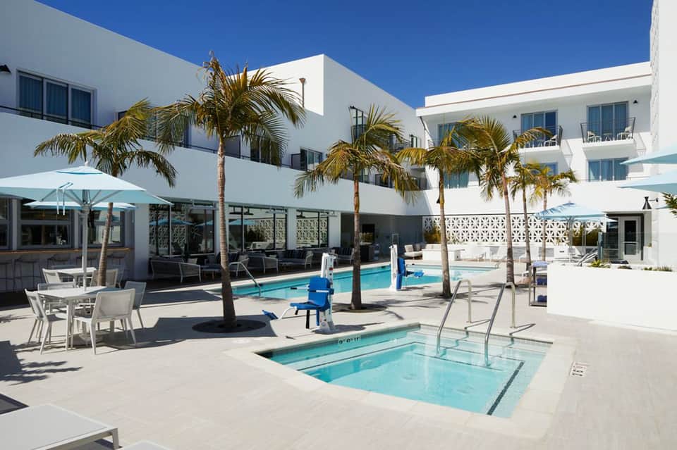 Contemporary resort pool area with white building, palm trees, loungers, and blue umbrellas