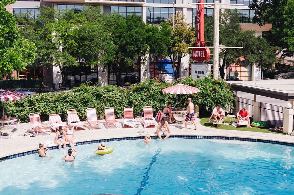 Retro-style pool with red and white striped loungers, guests swimming, and vintage motel sign visible
