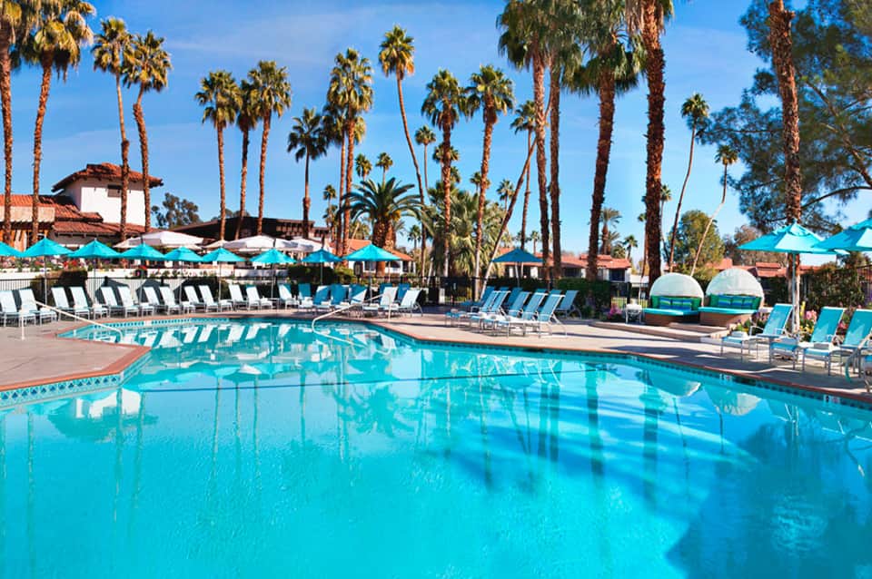 Resort swimming pool with turquoise water, palm trees, lounge chairs, and cabanas under blue sky