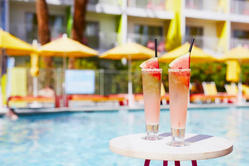 Two pink cocktails with straws by pool with yellow umbrellas and resort buildings