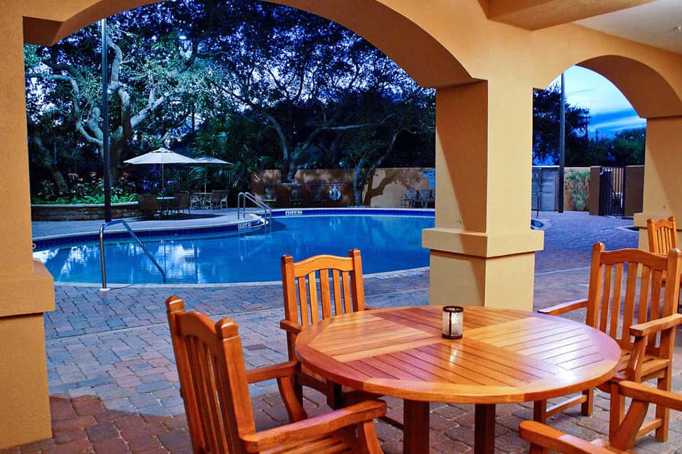 Covered pool patio with blue water, wooden dining table, orange chairs, and ocean view through arches