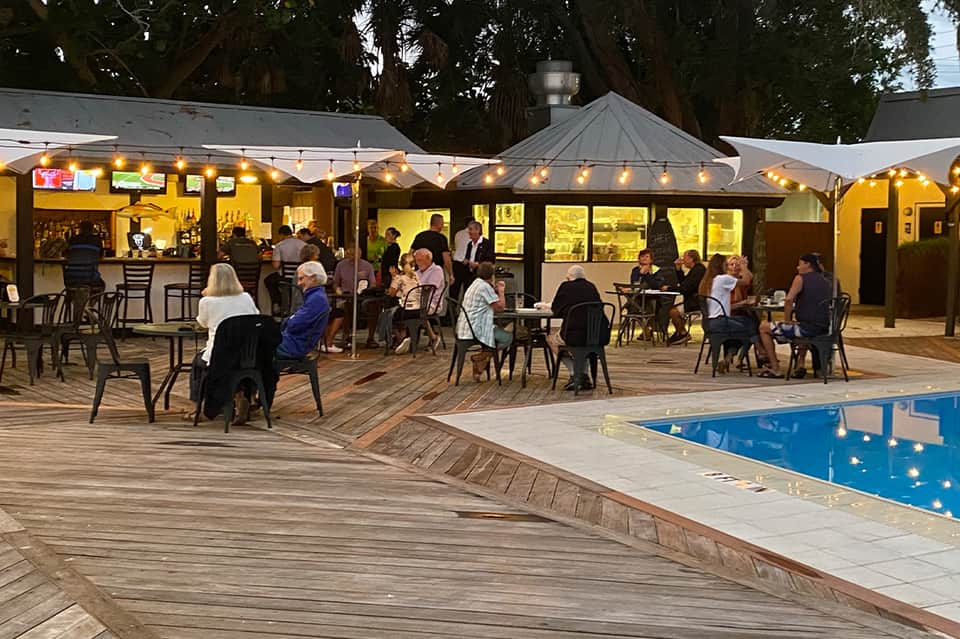 Evening resort pool deck with lit pavilions, bar area, and guests dining and socializing
