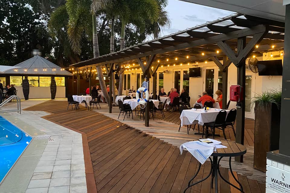 Outdoor dining patio with illuminated pergola, white tablecloths, and pool nearby at dusk