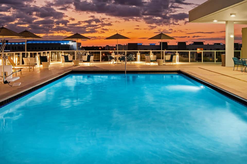 Resort rooftop pool at sunset with umbrellas, loungers, illuminated deck, and dramatic orange sky