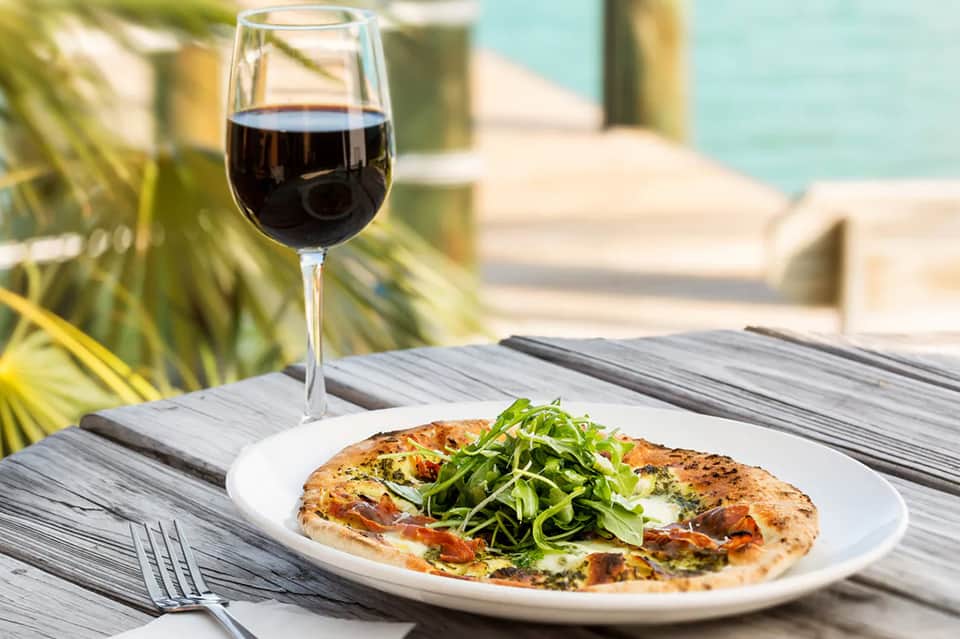 Gourmet pizza with wine glass on waterside table overlooking pool and tropical vegetation
