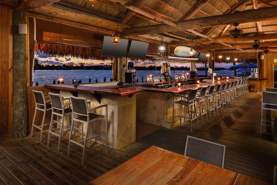 Waterfront tiki bar at sunset with wooden beams, long counter seating, and ocean view through open sides