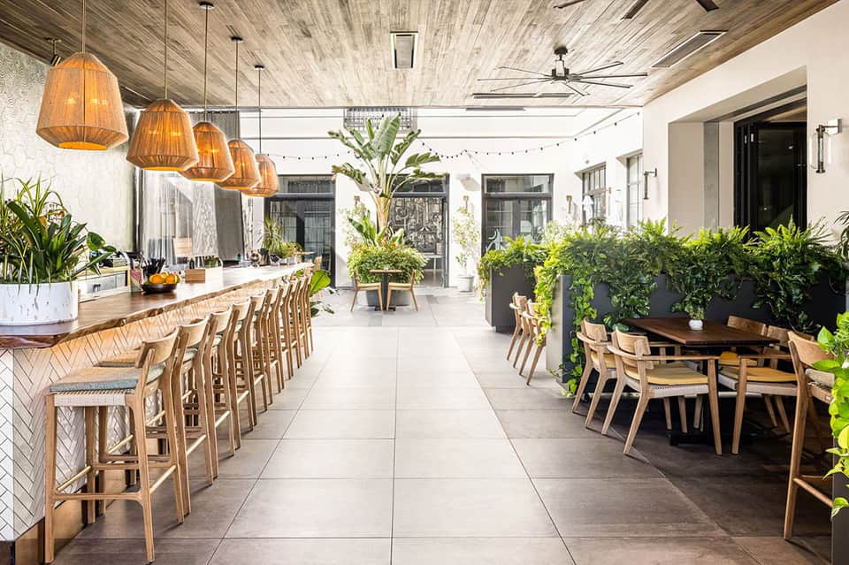 Modern open-air restaurant with wooden bar seating, pendant lights, potted plants, and natural light