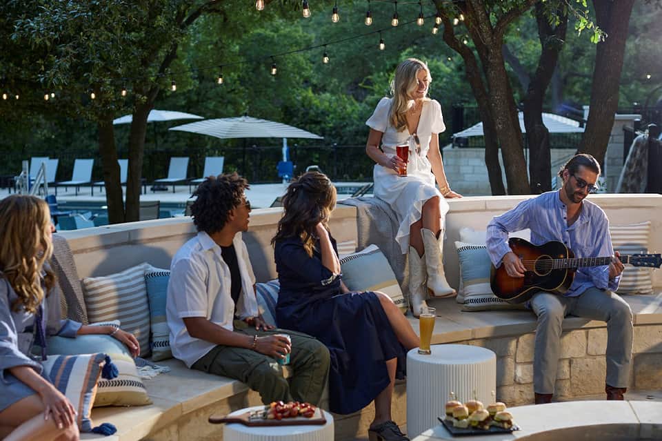 Guests relaxing on outdoor seating by pool with hanging lights, trees, and one playing guitar
