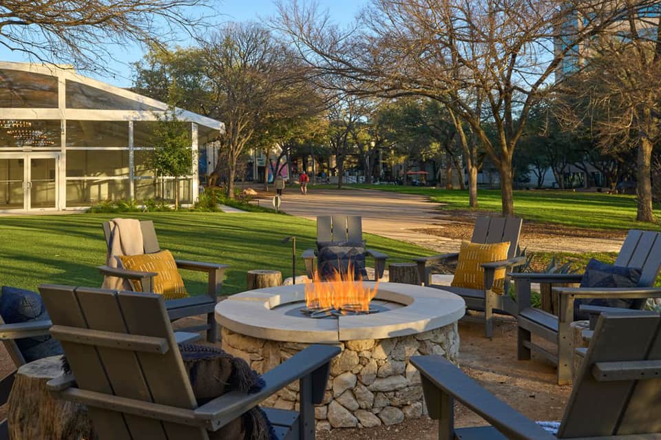 Outdoor fire pit seating area surrounded by trees with green lawn