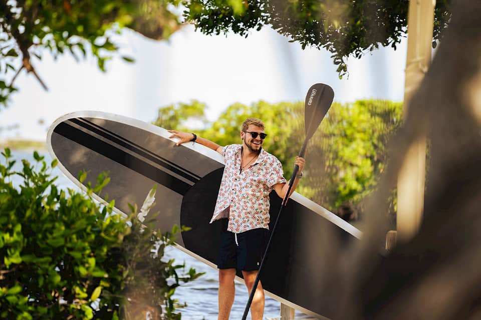 Man holding paddle board near water with tropical trees and mangroves