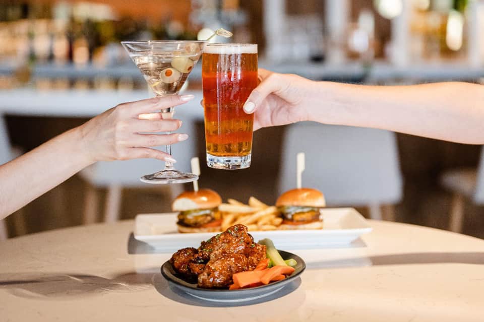 Two people toasting with cocktails at beachfront bar with food and drinks on table