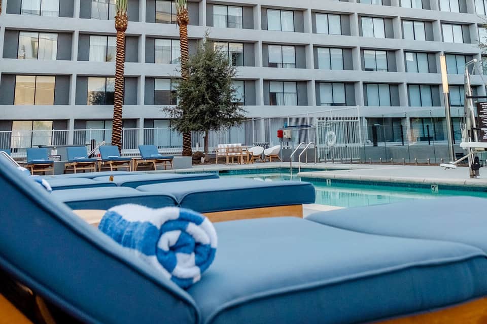 Modern hotel pool area with blue loungers, rolled towels, palm trees, and multi-story glass building backdrop