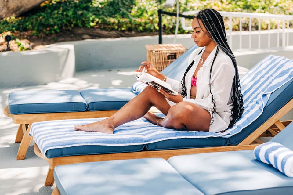 Woman reading on striped lounge chair in sunny resort pool area with hedges