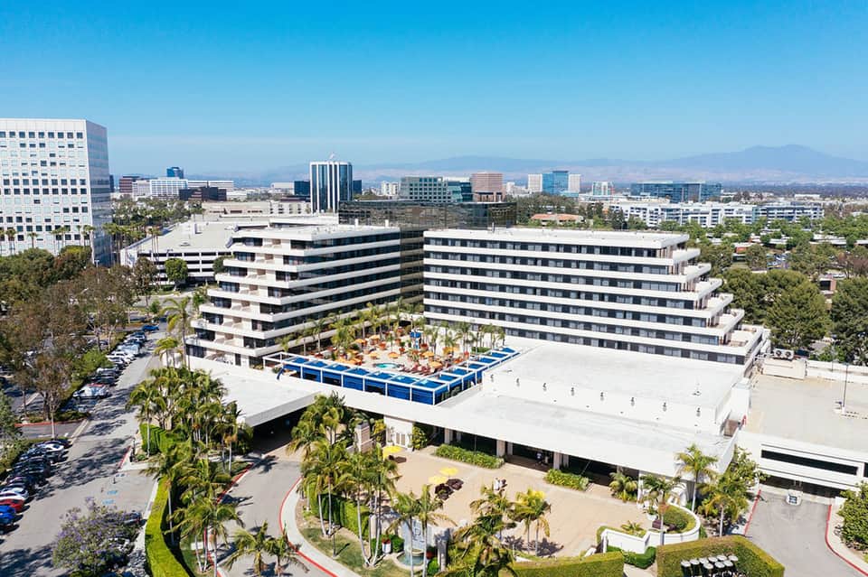 Modern curved hotel building with pool deck surrounded by palm trees and distant city skyline