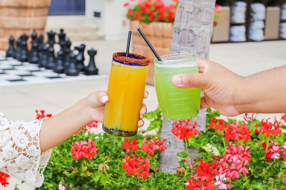 Two colorful tropical beverages with straws held outdoors near flowers