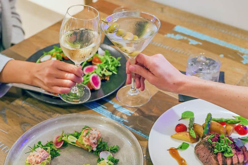 Two people toasting with wine glasses at dining table with gourmet plated dishes and fresh salad