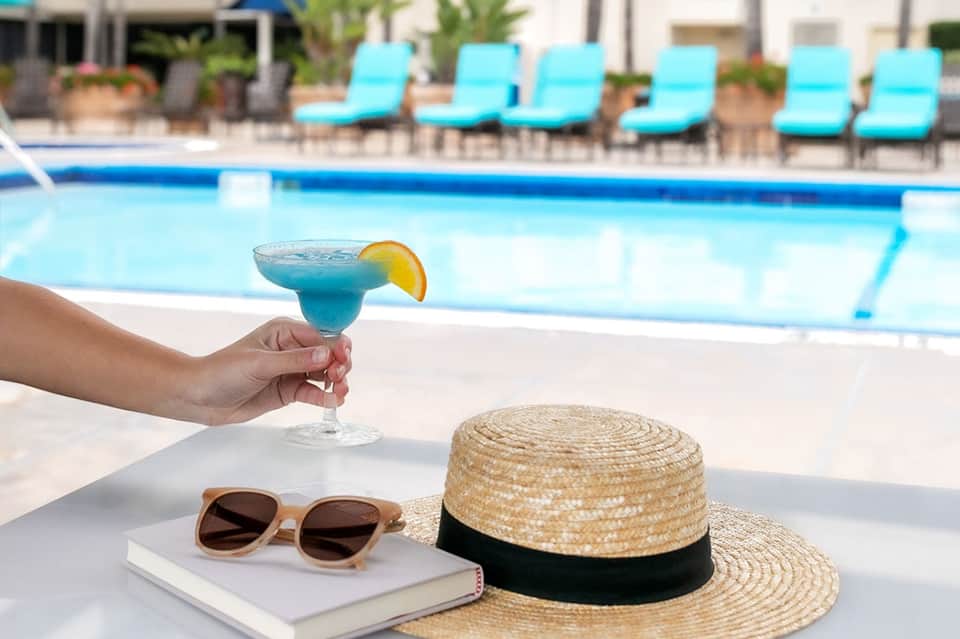 Blue cocktail with lemon slice held poolside with sunglasses, hat, and sunscreen on table