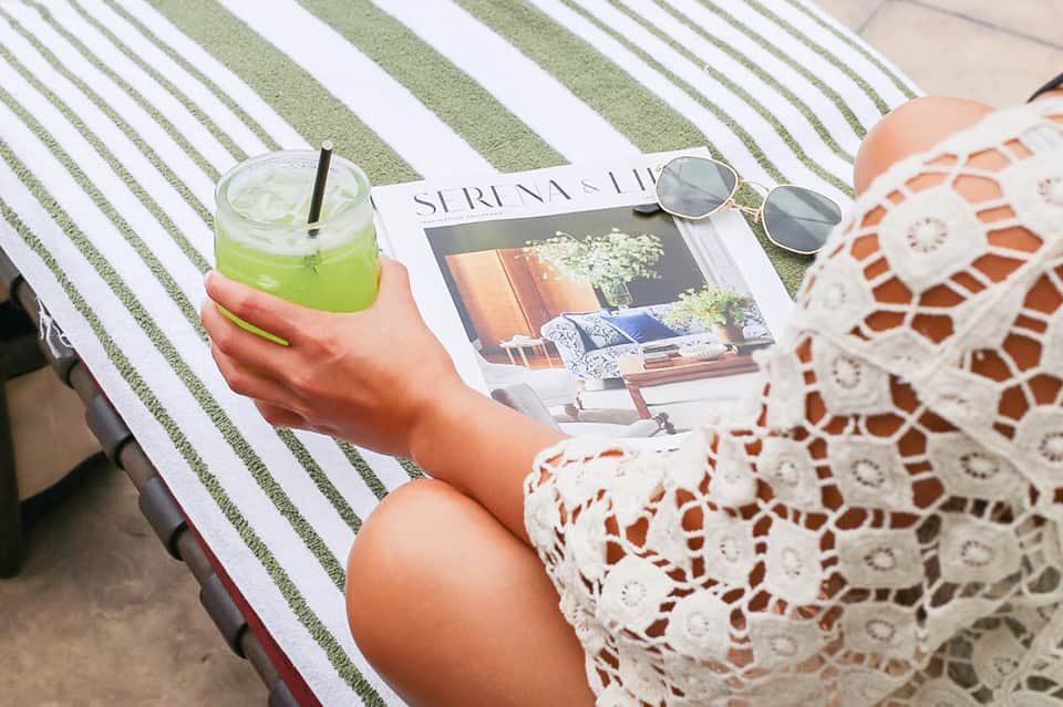 Woman relaxing poolside with green drink, magazine, sunglasses, and striped lounge chair
