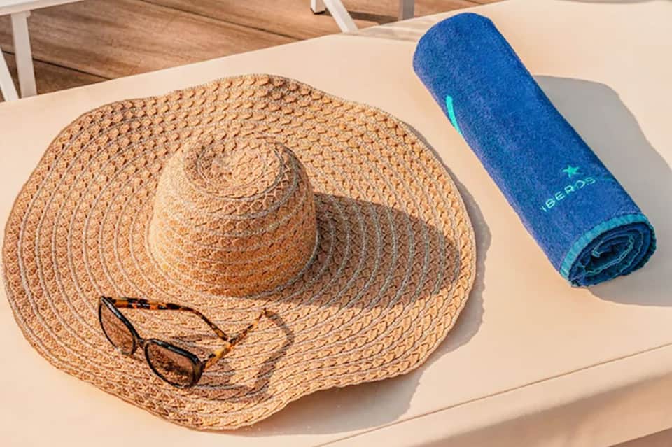Beach accessories on lounge chair including straw hat, sunglasses, and rolled blue towel in sunlight