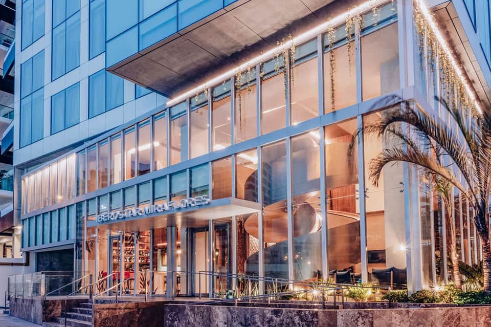 Modern restaurant with floor-to-ceiling windows, palm tree decor, and ambient evening lighting