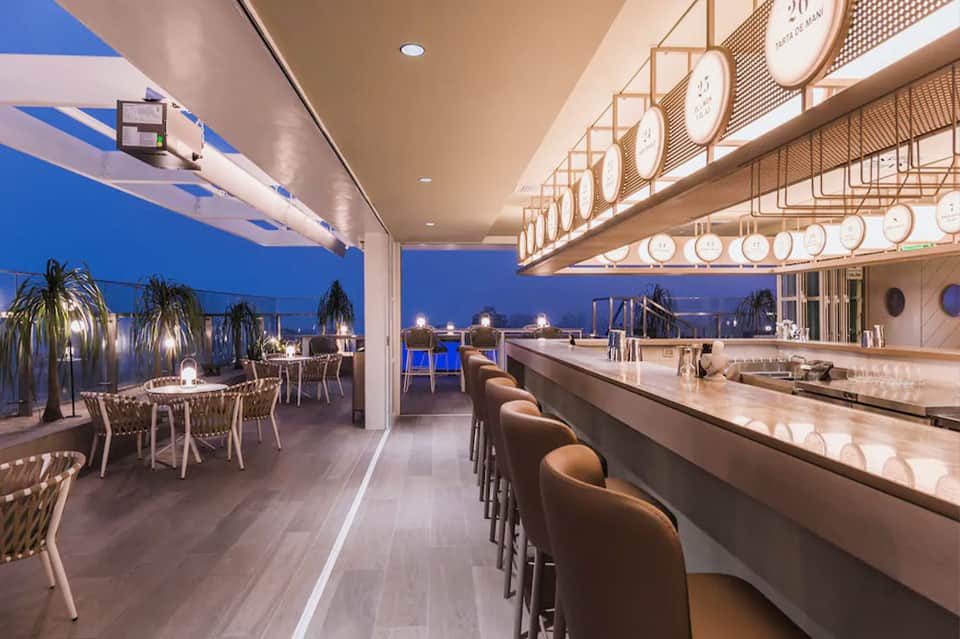 Upscale rooftop bar and restaurant with outdoor seating, blue water views, decorative plates, and illuminated evening setting