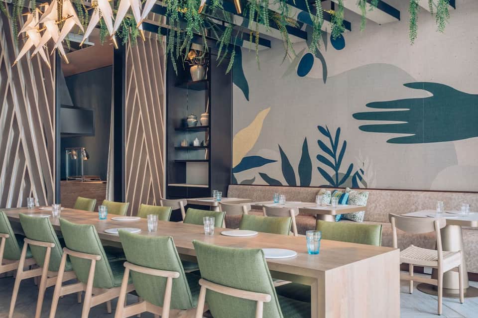Modern restaurant with botanical wall mural, wood tables, green chairs, and hanging plants